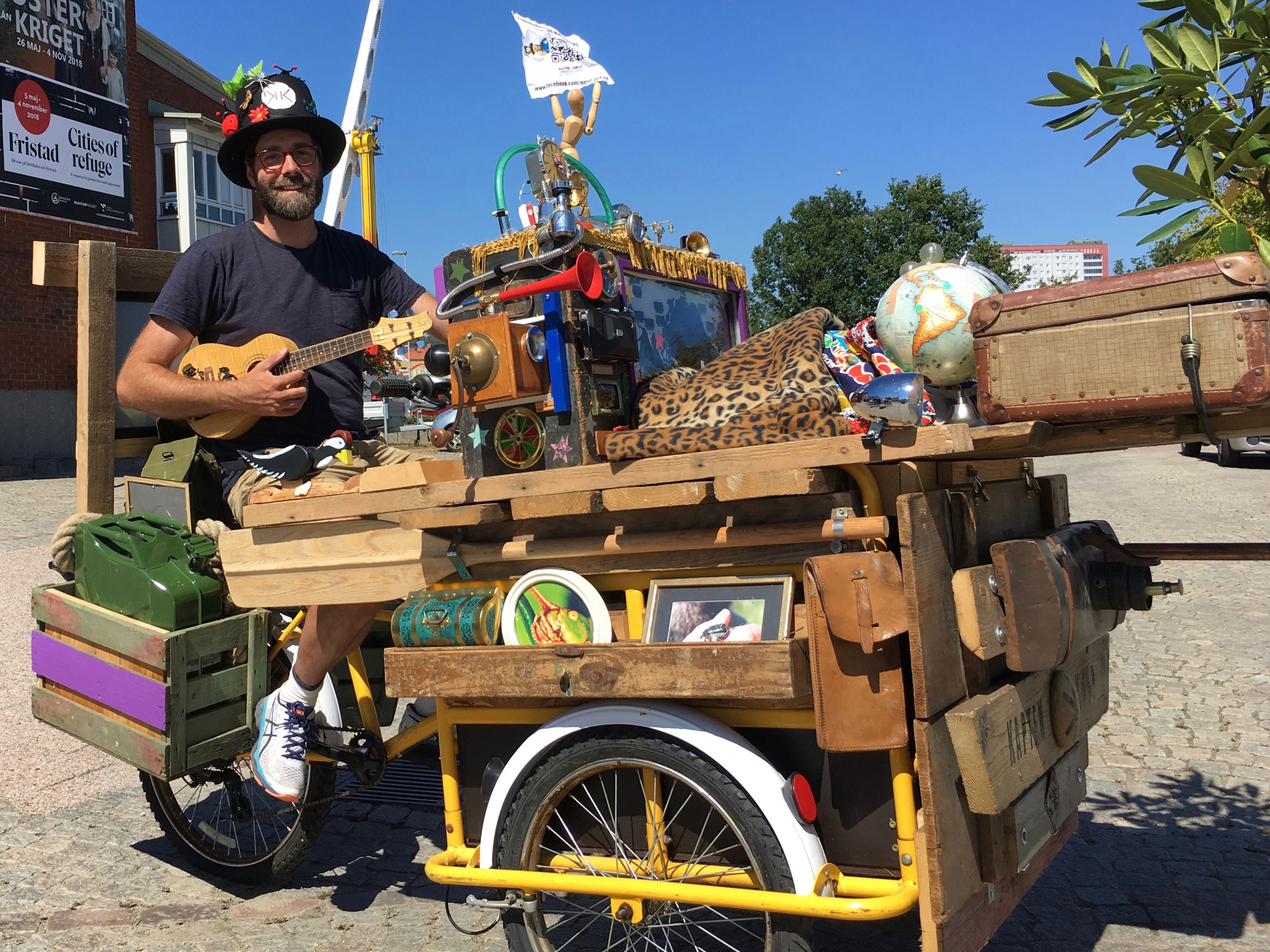 Bild på Kapten Kryp, som ler och håller i en ukulele - sittandes på en märklig cykel full av verktyg och manicker.