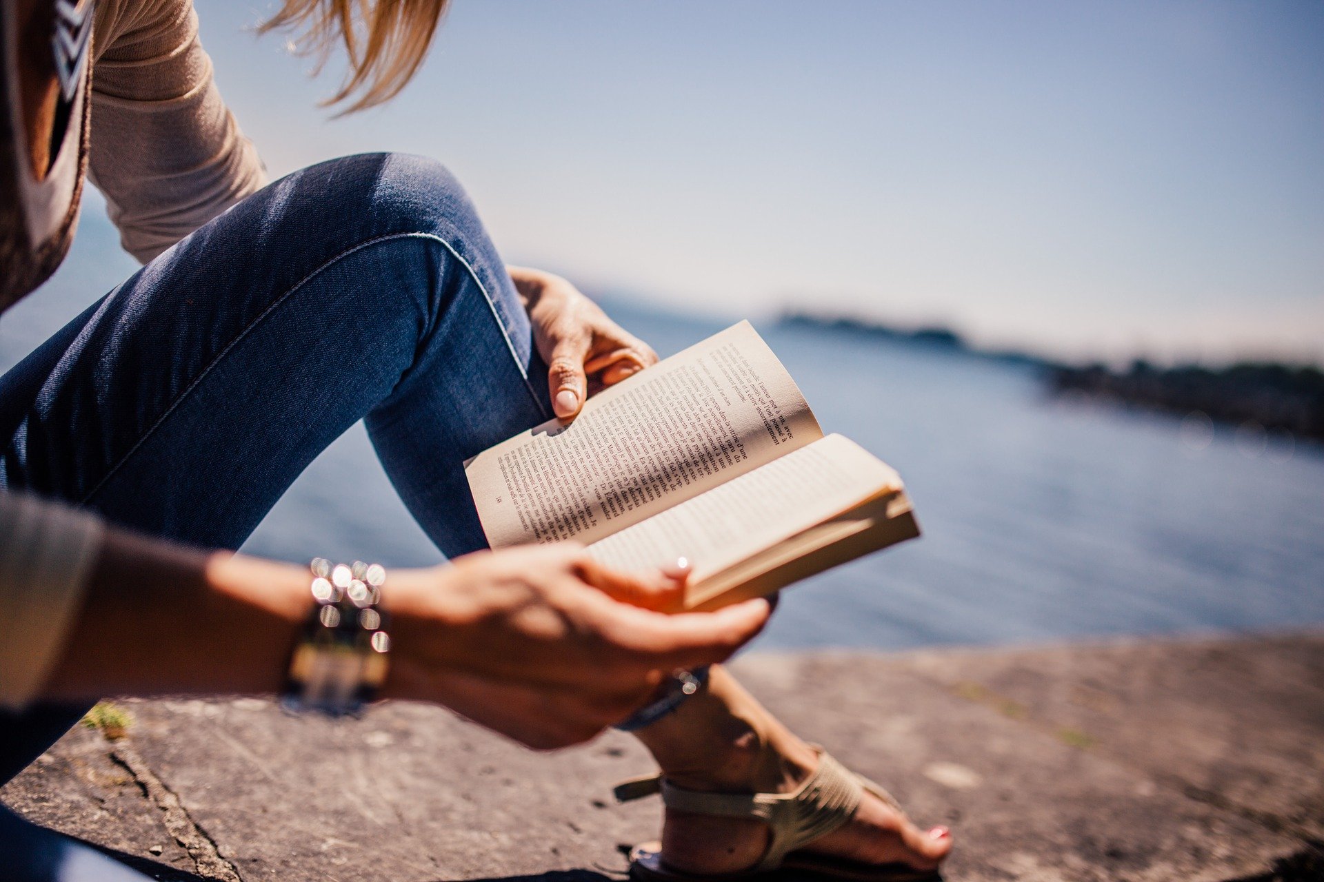 En kvinna sitter på en klippa vid vattnet och läser en bok.