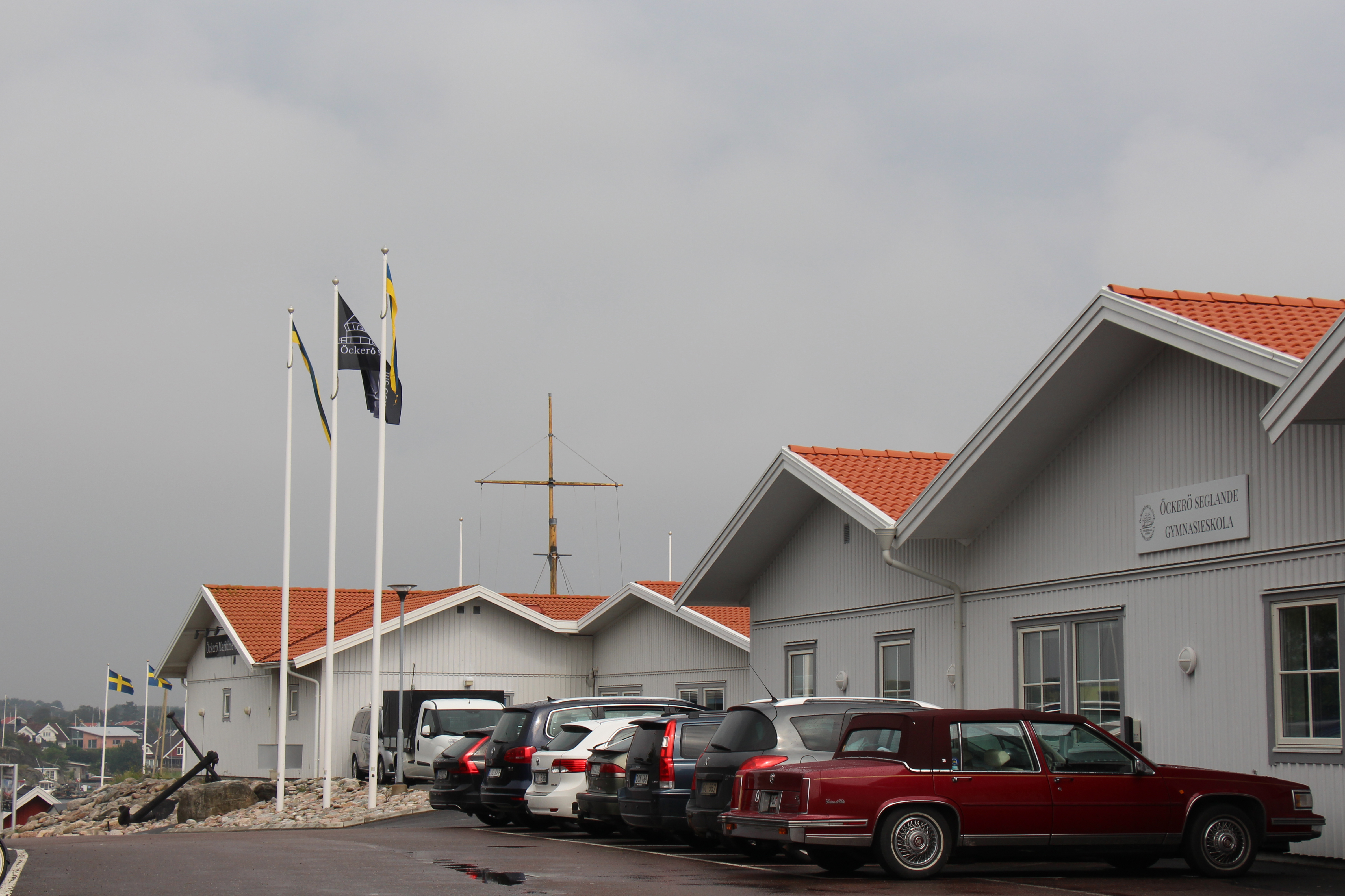 Bild på gymnasieskolans byggnad samt parkeringen med några bilar och flaggstänger.